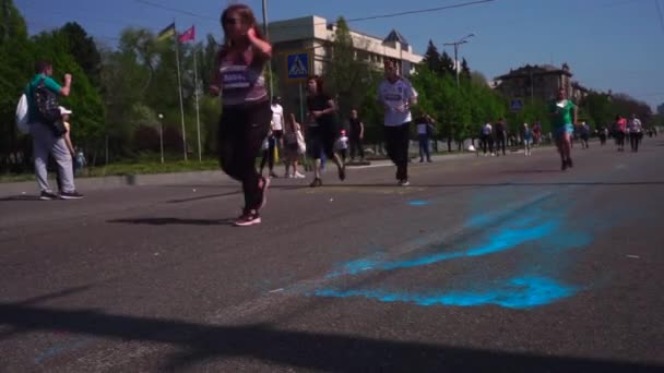 Wiele osób, dzieci biegać maraton wzdłuż drogi, wzdłuż alei, wzdłuż asfaltu, w farbie Holi, w różnych kolorach, w Zaporoże. Na asfalcie, Holi, żółty i niebieski. Szczęśliwy śmiech — Wideo stockowe