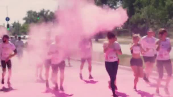 Os organizadores jogar, jogar pintura holi, rosa, cor, em face dos corredores de maratona que correm ao longo da avenida, na estrada de asfalto em camisetas brancas, bonés, óculos de sol e cobrir o nariz com o seu — Vídeo de Stock