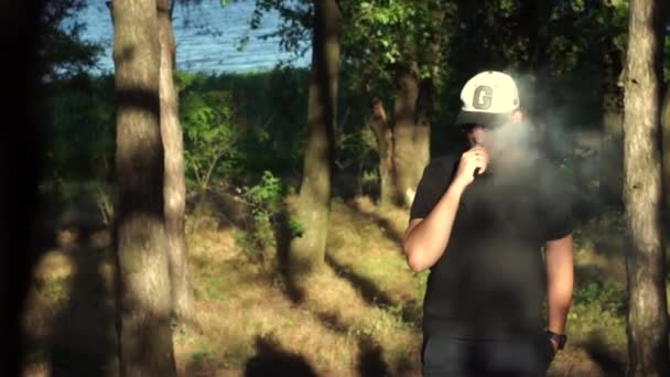 Tipo guapo y elegante en una gorra beige de moda con una visera negra y gafas, una camiseta y pantalones cortos, y un reloj con un toque de sin afeitar en el bosque, en la primavera, sobre el fondo de las cañas — Vídeo de stock