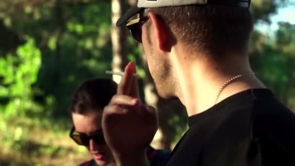 Gutaussehender, stylischer Typ mit beiger modischer Mütze mit schwarzem Visier und Sonnenbrille, T-Shirt und Shorts, Goldkette, mit einem Ring am Finger, mit einem Hauch von Unrasur im Wald, im Frühling — Stockvideo