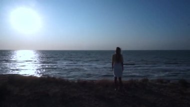 Kayalıkta, yeryüzünün kenarında, sarı çimenler üzerinde, sarı-beyaz elbiseli, uzun sarı saçlı, kameraya sırtını döneiç, başı eğik, maviye, yeşile bakan hareketsiz bir kız duruyor.