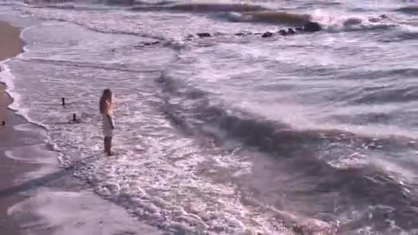 Uma menina bonita e triste com cabelos longos e loiros em um vestido amarelo-branco caminha na areia, costa, praia e caminha para o mar lamacento. Vento, ondas, sol, pedras negras, verão, quente, ventoso, dia, espuma, surf . — Vídeo de Stock