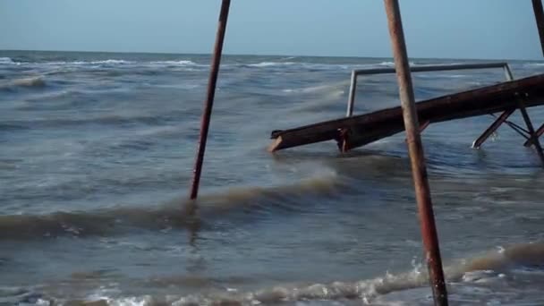 Pavillons anciens, cassés, détruits, auvents, inondés d'eau sur la plage. Petites vagues de la mer boueuse, toit coulé, fondations, côte, sable jaune, pierres noires contre un ciel bleu brillant, avec un — Video