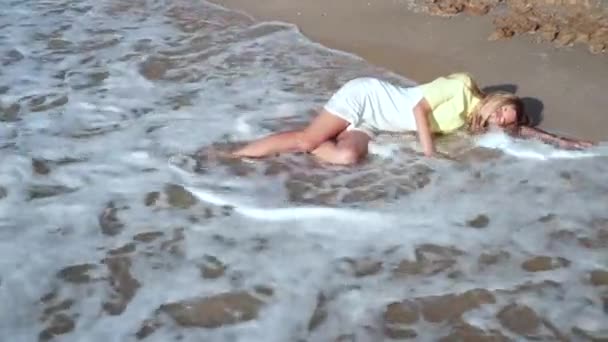 黄色白いドレスと長いブロンドの髪を着た美しい、悲しい少女は、海岸で目を閉じて、波で覆われ、泡で覆われ、彼女の顔を覆っています. — ストック動画