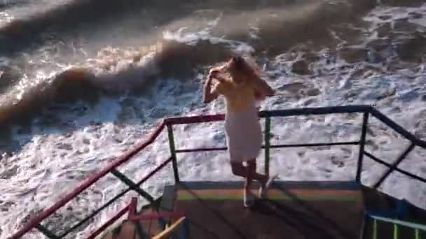 Een mooi meisje met lang haar, een geel-witte jurk staat en recht haar haar op het uitkijkplatform, stappen, met haar terug naar de zee, leunend op de multi-gekleurde leuningen en op zoek — Stockvideo