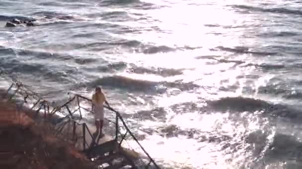 Krásná dívka s dlouhými vlasy, žlutými a bílými šaty stojí na pozorovací palubě, schody a hledí na moře, opřená o mnohobarevné zábradlí. Vítr, vlny, modrá obloha, sluneční dráha. — Stock video