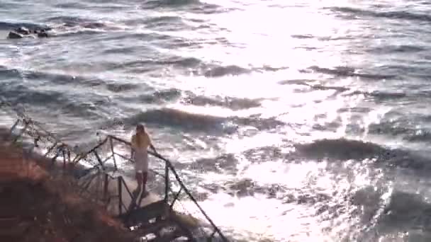 Una hermosa chica con el pelo largo, vestido amarillo y blanco se encuentra en la plataforma de observación, pasos, y mira al mar, apoyándose en las barandillas multicolores. Viento, olas, cielo azul, camino solar . — Vídeos de Stock