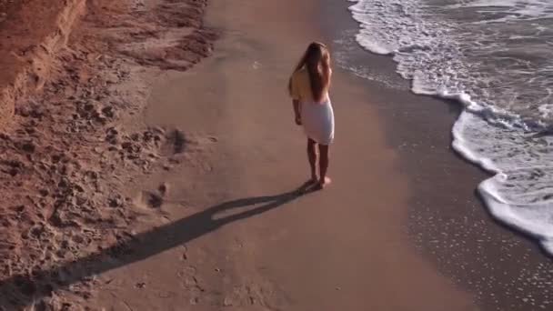 Uma menina bonita com cabelos longos, em um vestido amarelo-branco, caminha descalça na areia amarela, praia, costa, mar. No final, a menina vira-se. À esquerda está uma rocha laranja, uma montanha de barro, sobre — Vídeo de Stock