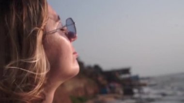 Uzun sarı saçlı, kırmızı tişörtlü, sıra dışı, dikdörtgen, mor gözlüklü güzel bir kız gözleri kapalı güneşe bakar ve mavi denize, sahile, sahile karşı sıcak ışınlarda güneşe bakar.