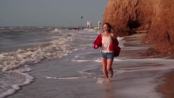 Une belle fille aux longs cheveux blonds, vêtue d'une veste rouge, avec des lunettes violettes rectangulaires insolites, une chaîne, un short en denim, les pieds nus, un T-shirt rayé court le long du rivage, la plage, du sable mouillé — Video