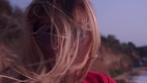 Красивая девушка с длинными светлыми волосами, в красной футболке, в необычных, прямоугольных, фиолетовых очках смотрит в камеру на фоне моря, берега, пляжа и гор — стоковое видео