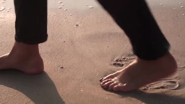 Os pés descalços da menina, em jeans pretos, saem, avançando, deixando marcas de calcanhar, uma onda com espuma sobe e lava os trilhos na areia úmida e amarela da praia . — Vídeo de Stock