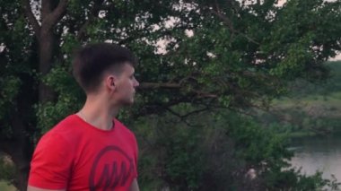 Kırmızı t-shirt bir zincir ile düzgün bir adam yeşil, güzel bir ağaç, diğer tarafı, nehir, doğa, çim bakmak