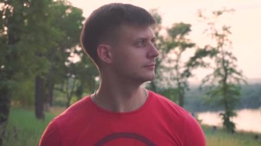 kırmızı t-shirt yakışıklı adam yan bakar, kameraya gülümseme, yeşil ağaçların arka planına karşı, çimen, yaprakları