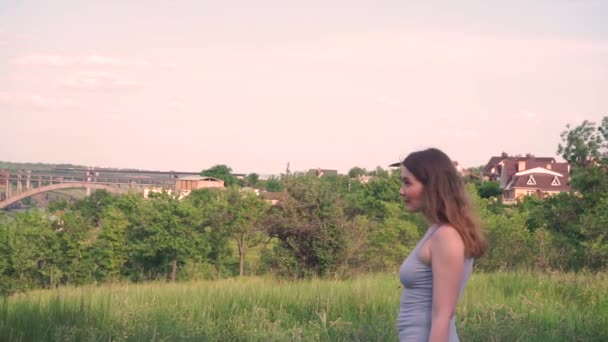 Belle femme va, regarde vers l'avant sur le fond d'arbres verts, herbe, rivière, pont, village, maisons, nature aux cheveux blonds — Video