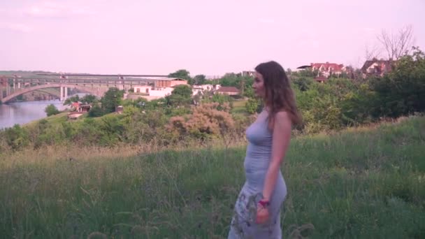 Молода жінка йде і дивиться вперед на тлі зелених дерев, трави, річки, мосту, села, будинків, природи з світлим волоссям — стокове відео