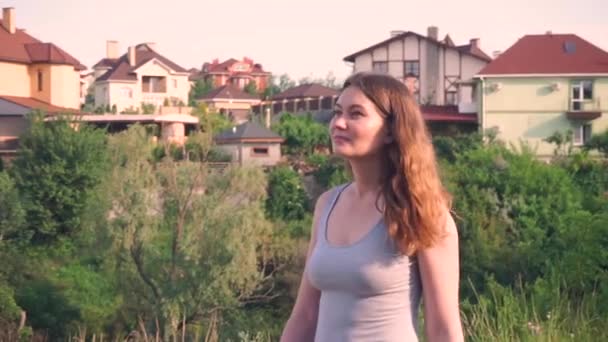 Menina bonita, olha para a frente no fundo de árvores verdes, casas, natureza, cabelo loiro vai — Vídeo de Stock