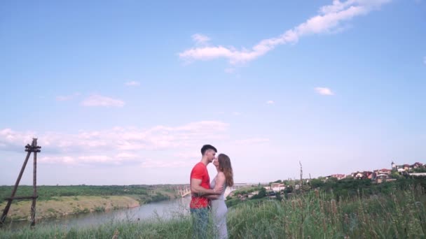 Plan général, beau baiser homme et femme au centre du cadre, pont, rivière, arbres, herbe, maisons sur fond — Video