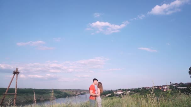 Plan général, beau baiser homme et femme au centre du cadre, pont, rivière, arbres, herbe, maisons sur fond — Video