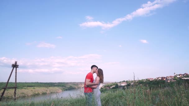 Algemeen plan, knappe man en vrouw na een kus lachen en gek rond in het midden van het frame. brug, rivier, bomen, gras, huizen tegen de achtergrond — Stockvideo