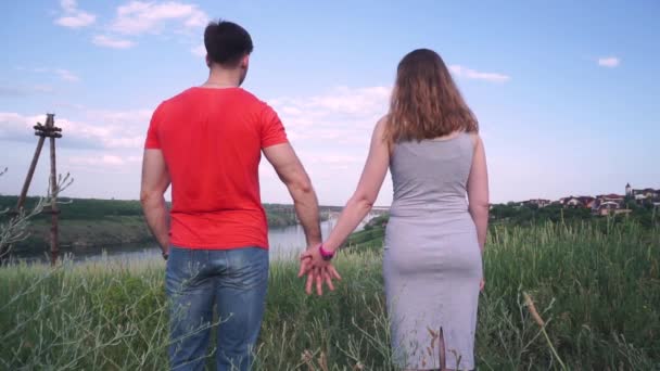 Молодой, красивый парень и девушка держатся за руки и идут вперед, на фоне моста, реки, дерева, неба — стоковое видео