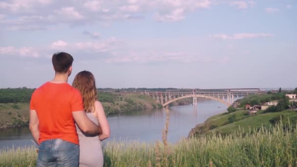 Schöner Kerl, der Mädchen von hinten umarmt und beide verneigen sich vor der Natur, Brücke. Gesamtplan — Stockvideo