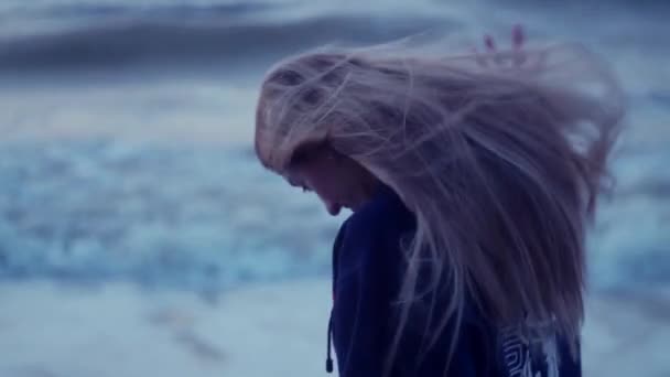 Mooi meisje kijkt naar de zee en draait om de camera, haar haar ontwikkelt, wind, golven tegen de achtergrond — Stockvideo