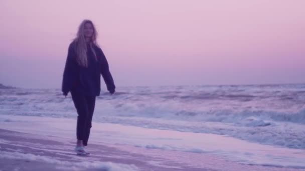 Menina com belos passeios de cabelo ao amanhecer ao longo da areia, na costa junto ao mar. Céu rosa, ondas, vento no fundo — Vídeo de Stock