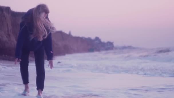 Het meisje loopt op het natte zand van de zee bij zonsopgang en raakt het schuim van de golven — Stockvideo