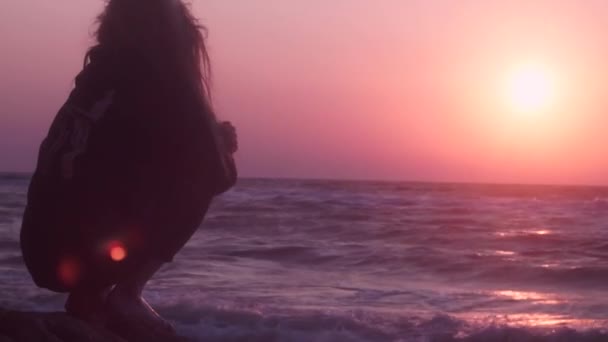 Mooi meisje kijken roze zonsondergang op de zee van donkere kleur zittend op een steen — Stockvideo