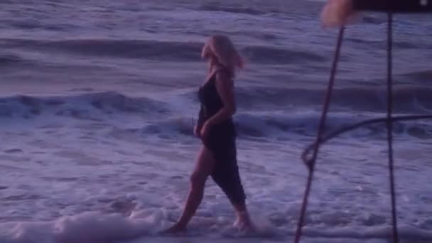 Uma mulher de vestido preto caminha na praia, na água do mar, ondas, espuma — Vídeo de Stock