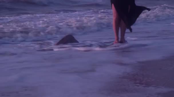 Πόδια μιας γυναίκας με ένα μαύρο φόρεμα, σε μια πέτρα, στη θάλασσα, κατά τη διάρκεια του σερφ, κύματα — Αρχείο Βίντεο