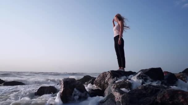 Mädchen im T-Shirt steht auf Felsen im Meer, blickt auf Horizont, um Wellen herum, plantscht — Stockvideo