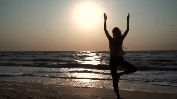 Chica de pie en una pose de yoga, frente al sol al atardecer, en la playa, en el mar — Vídeos de Stock