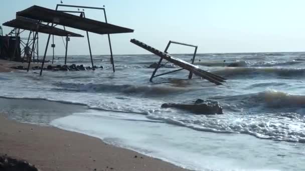 Pabellones viejos, rotos, destruidos, toldos, inundados de agua en la playa. Pequeñas olas del mar fangoso, techo vertido, cimientos, costa, arena amarilla, piedras negras contra un cielo azul brillante, con un — Vídeos de Stock