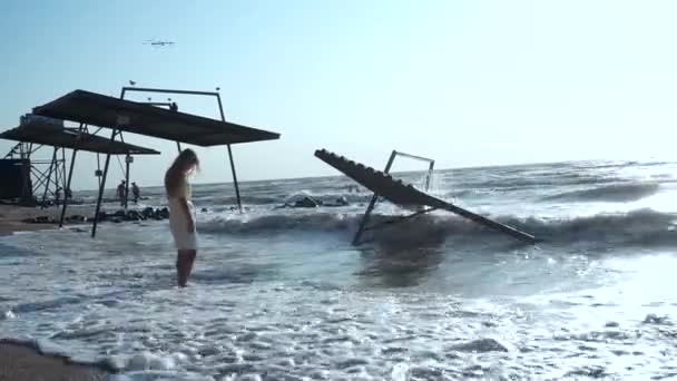 Beautiful girl bowing her head, stands in the waves of the sea, on the beach, near the pavilions — Stock Video