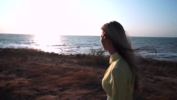 Молодая девушка идет против ветра вдоль берега моря, смотрит на солнце в голубом небе — стоковое видео