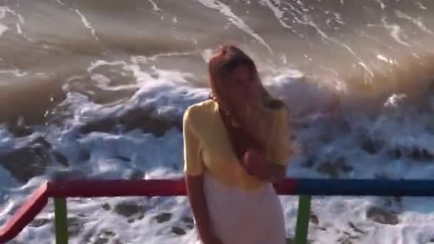 Câmera se afasta de uma linda garota apoiada em um corrimão multicolorido, contra o fundo do mar — Vídeo de Stock