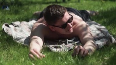 Siyah gözlüklü adam çimlere, paspaslara uzanır, kameraya bakar, gülümser, şakalar yapar, güler.