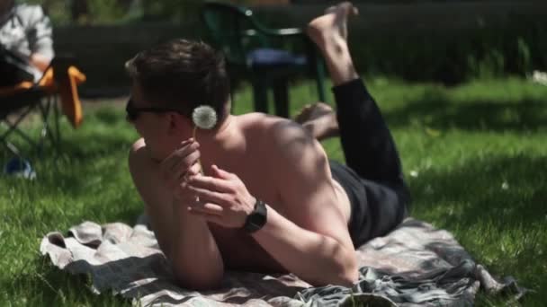 Guy sunglasses, lying grass, litter, sniffs yellow dandelion summer cottage — Stock Video