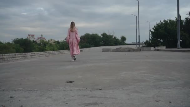 Uma menina caminha ao longo de uma estrada de concreto deserta, uma ponte inacabada — Vídeo de Stock