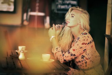 Sarışın bir kadın öğle yemeği molasında açık hava kafeteryasında oturuyor. Yanında kahve fotografı var.