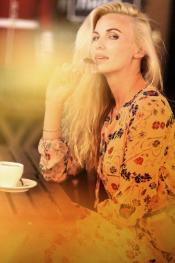 Sarışın bir kadın öğle yemeği molasında açık hava kafeteryasında oturuyor. Yanında kahve fotografı var.