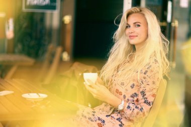 Sarışın bir kadın öğle yemeği molasında açık hava kafeteryasında oturuyor. Yanında kahve fotografı var.