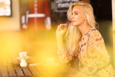 Sarışın bir kadın öğle yemeği molasında açık hava kafeteryasında oturuyor. Yanında kahve fotografı var.