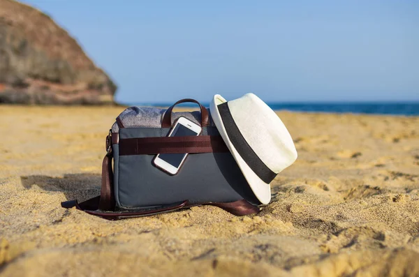 Shoulder bag with a smartphone and a white hat on the sand of the beach