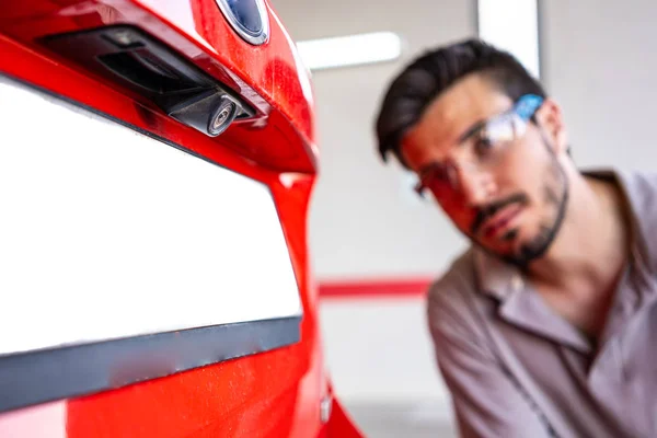 Bir araç muayene atölyesinde bir arabanın arka görüş kamerasını inceleyen güvenlik gözlüklü teknisyen