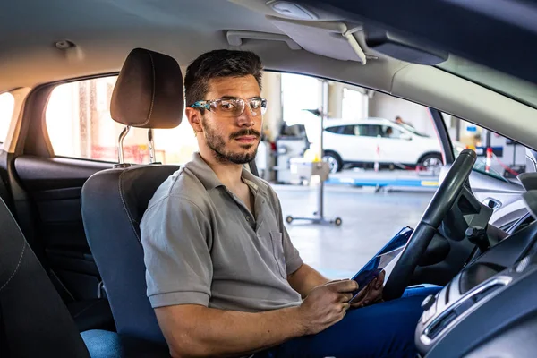 Bir araç muayenesi sırasında bir liste kontrol kamera bakarak bir araba koltuğunda oturan güvenlik gözlükleri ile Teknisyen
