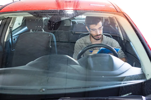 Technician with safety glasses sitting in a car seat checking a list during a vehicle inspection