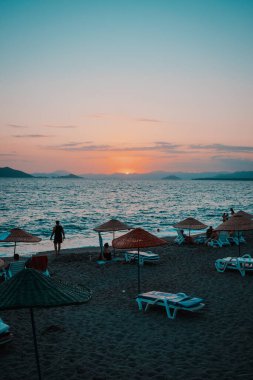 Güzel bir plajın dikey bir resmi. Önünde bir gün batımı, arkasında da bir siluet var.